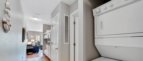 Une buanderie avec un sèche-linge blanc et un lave-linge fixé au mur. Un couloir, orné d’un tapis à motifs bleus, mène à un espace de vie comprenant un canapé et une table basse.