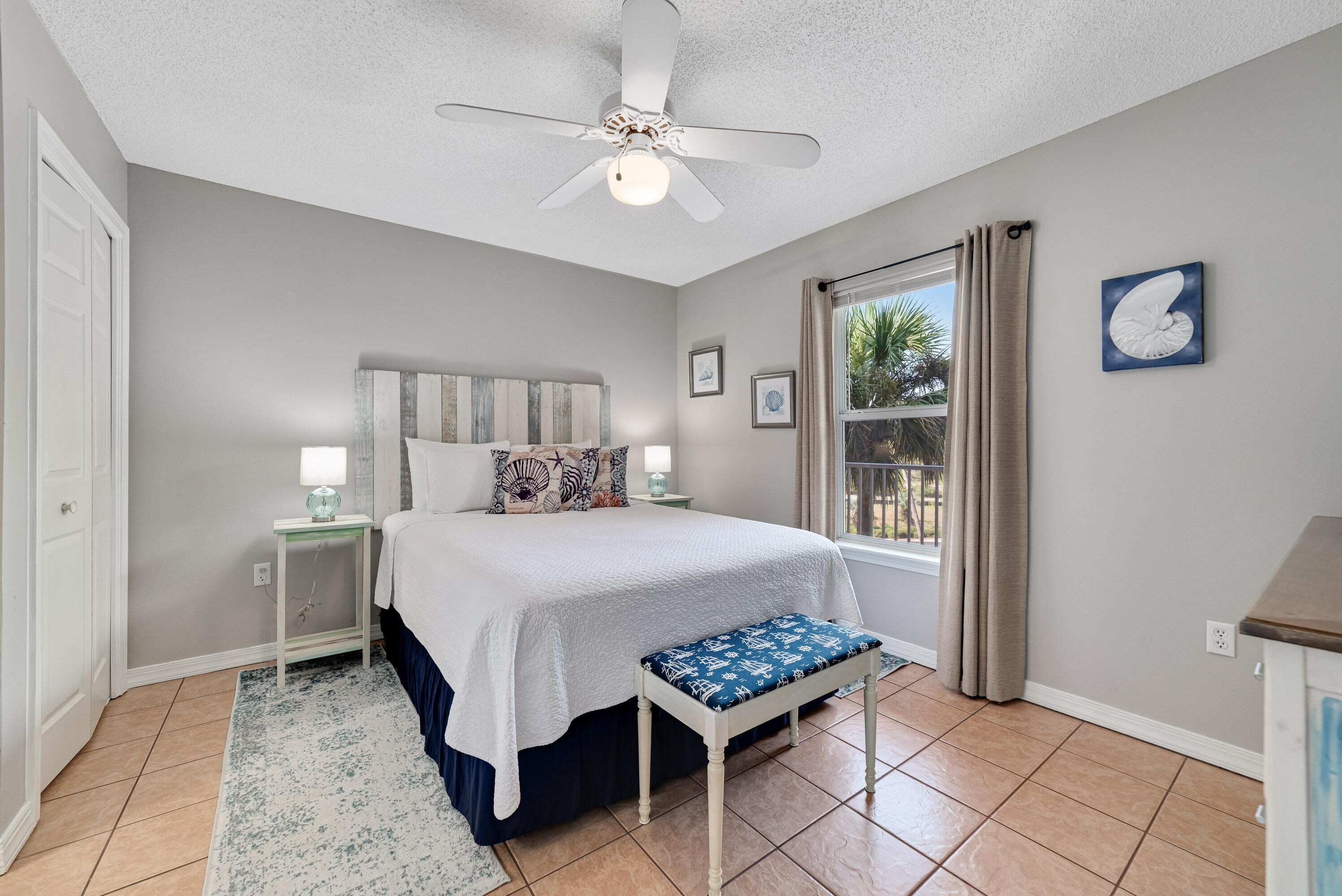 A bedroom with a bed, a nightstand, a bench, a ceiling fan, a window with curtains, and framed pictures on the wall.