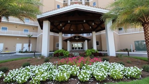 A hotel entrance with a covered patio, palm trees, and flower beds. - Vista Del Mar 406 (Pensacola)