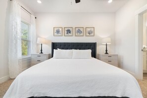 A neatly arranged bedroom with a large bed, two bedside tables with lamps, and a window with sheer curtains. - Blue Angel Casa (Pensacola)