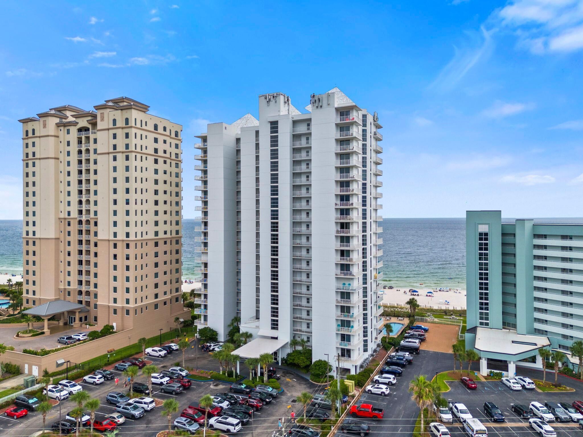 A high-rise hotel with multiple floors, a parking lot, and a beach view.