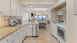 A kitchen with white cabinets, a granite countertop, a dishwasher, a washing machine, and a dining area with a chandelier. - Shipwatch D401 (Pensacola)