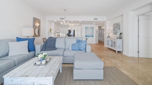 A modern living room with a grey sofa, a coffee table, and a dining area with a white cabinet and a chandelier. - Indigo East 1502 (Pensacola)