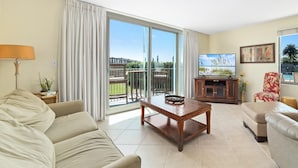 A living room with a sofa, a coffee table, a TV, and a view of a balcony with outdoor furniture.