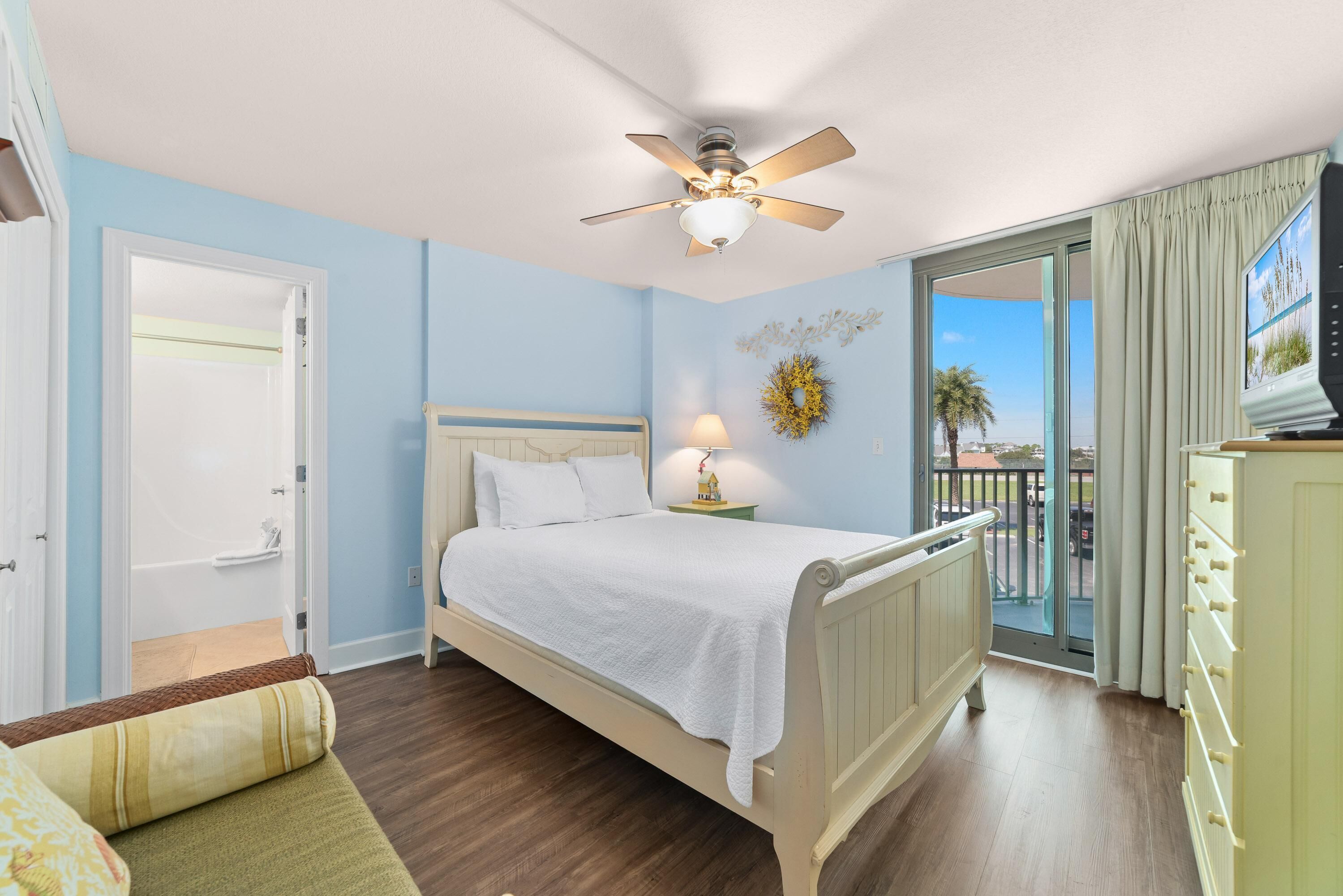 A bedroom with a bed, a ceiling fan, a nightstand with a lamp, a view of the outdoors, and a television.