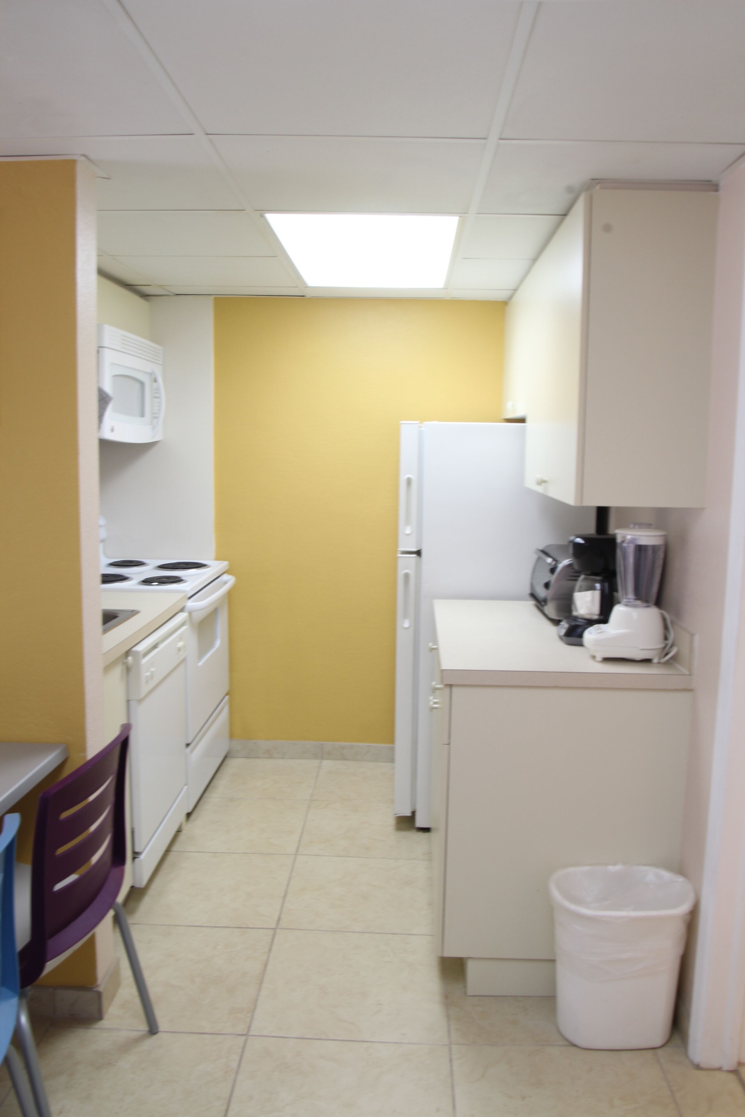 A compact kitchen with white appliances, a microwave, stove, refrigerator, and a trash bin.