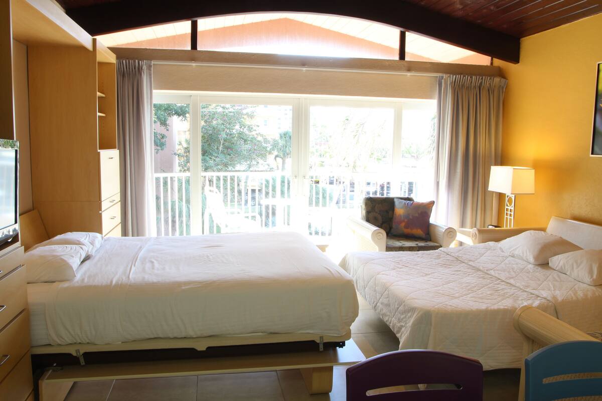 A hotel room with two beds, a television, a chair, and a balcony with curtains.