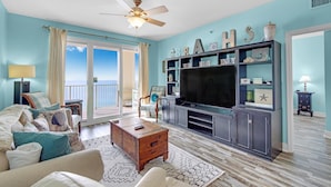 A living room with a large TV, a ceiling fan, and a view of the ocean through the sliding glass doors. - Ariel Dunes I 2202 (Miramar Beach)