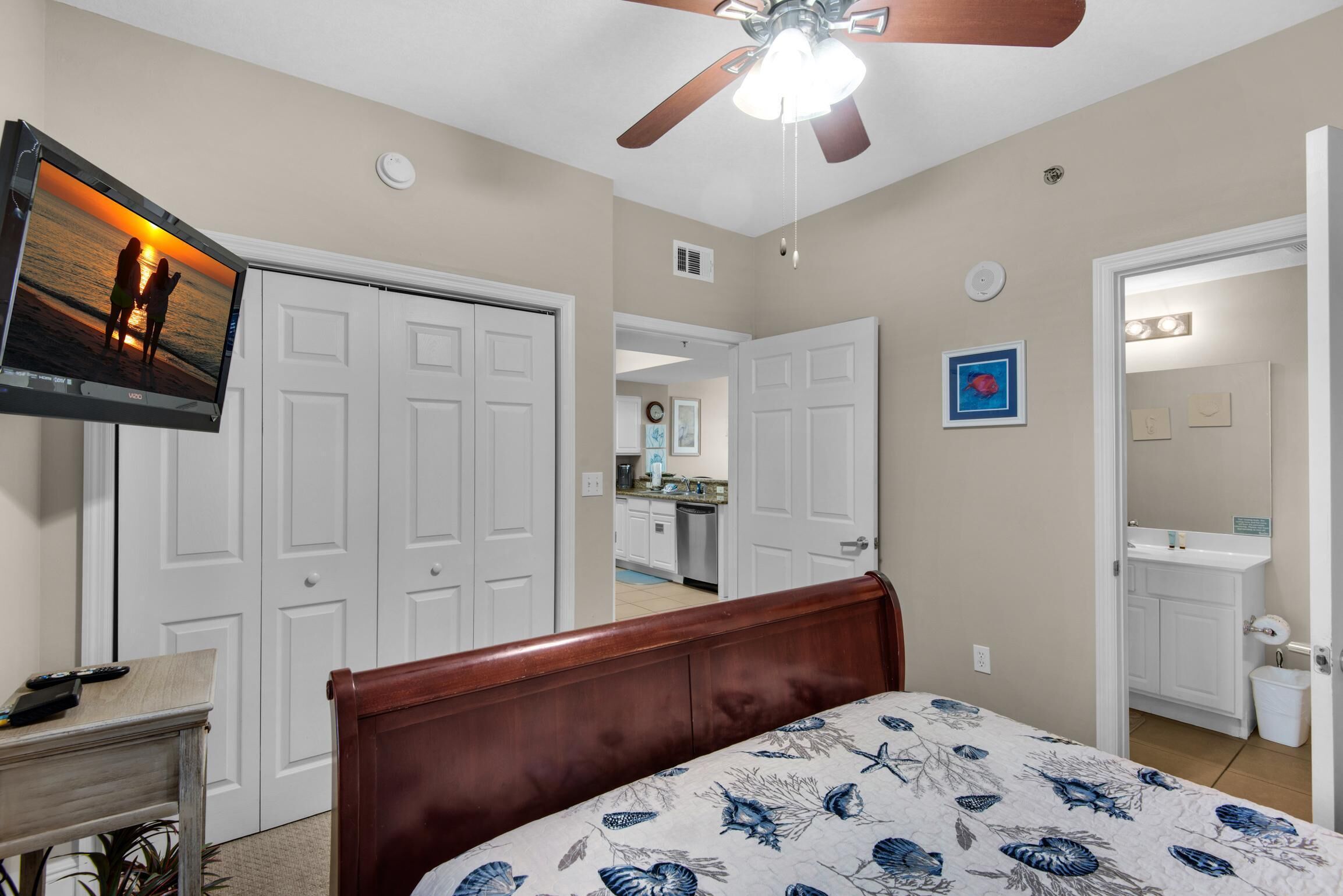 A bedroom with a bed, a TV mounted on the wall, a ceiling fan, and a view into the adjacent kitchen.
