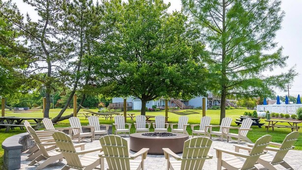 A circular outdoor seating area with Adirondack chairs around a central fire pit, set on a paved patio with a grassy lawn and trees in the background. - Waterfront in Historic Rock Hall, overlooking the main harbor and Chesapeake Bay (Rock Hall)