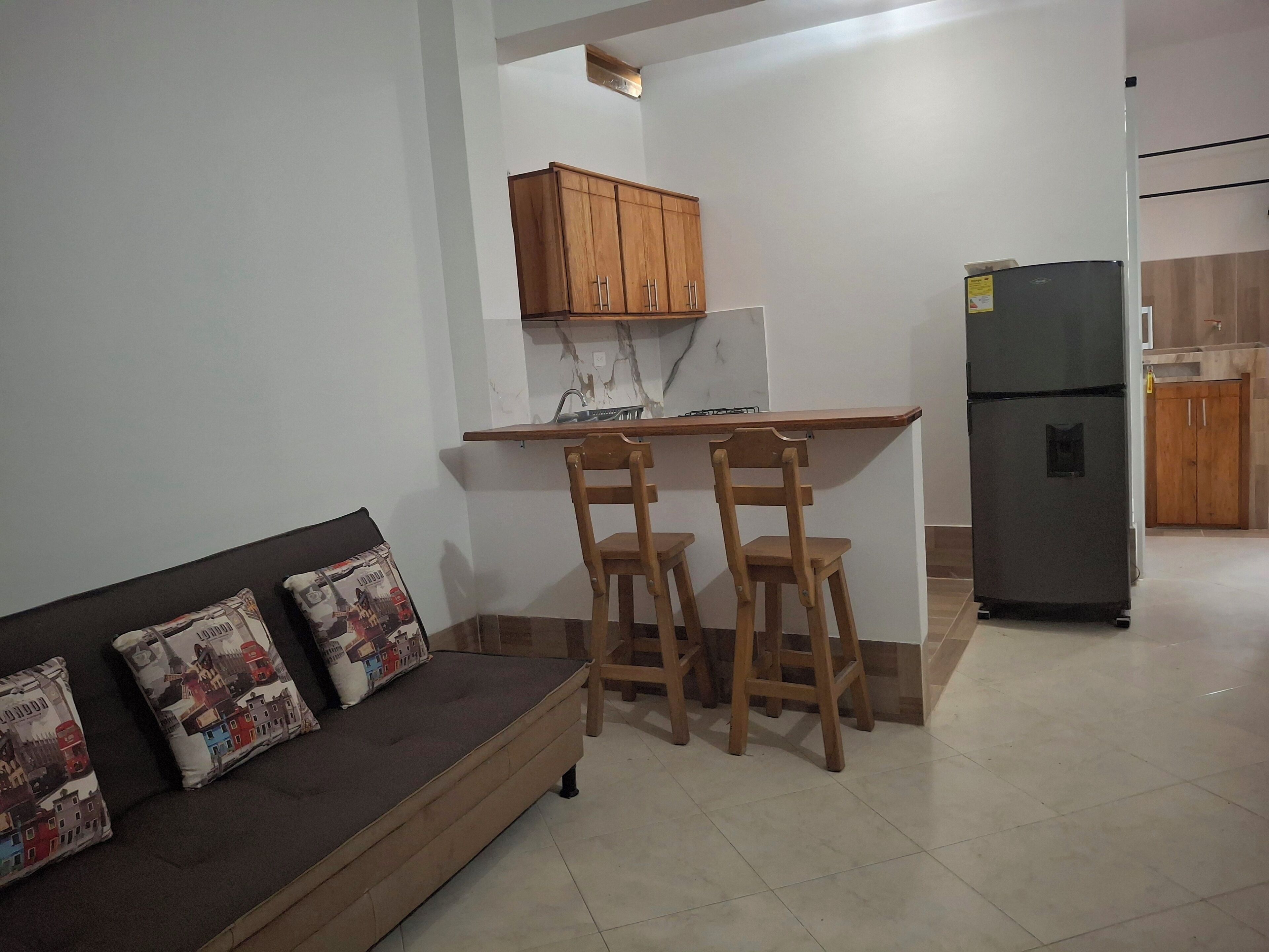Een compacte keuken met houten kastjes, een ingebouwde gootsteen en een koelkast. Er is een klein eethoekje met twee houten stoelen en een bankje.