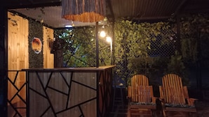 An outdoor bar area with a wooden counter, a decorative wall with a palm tree and sunset design, and two wooden chairs. - CABAÑAS MAR CARIBE (Necoclí)