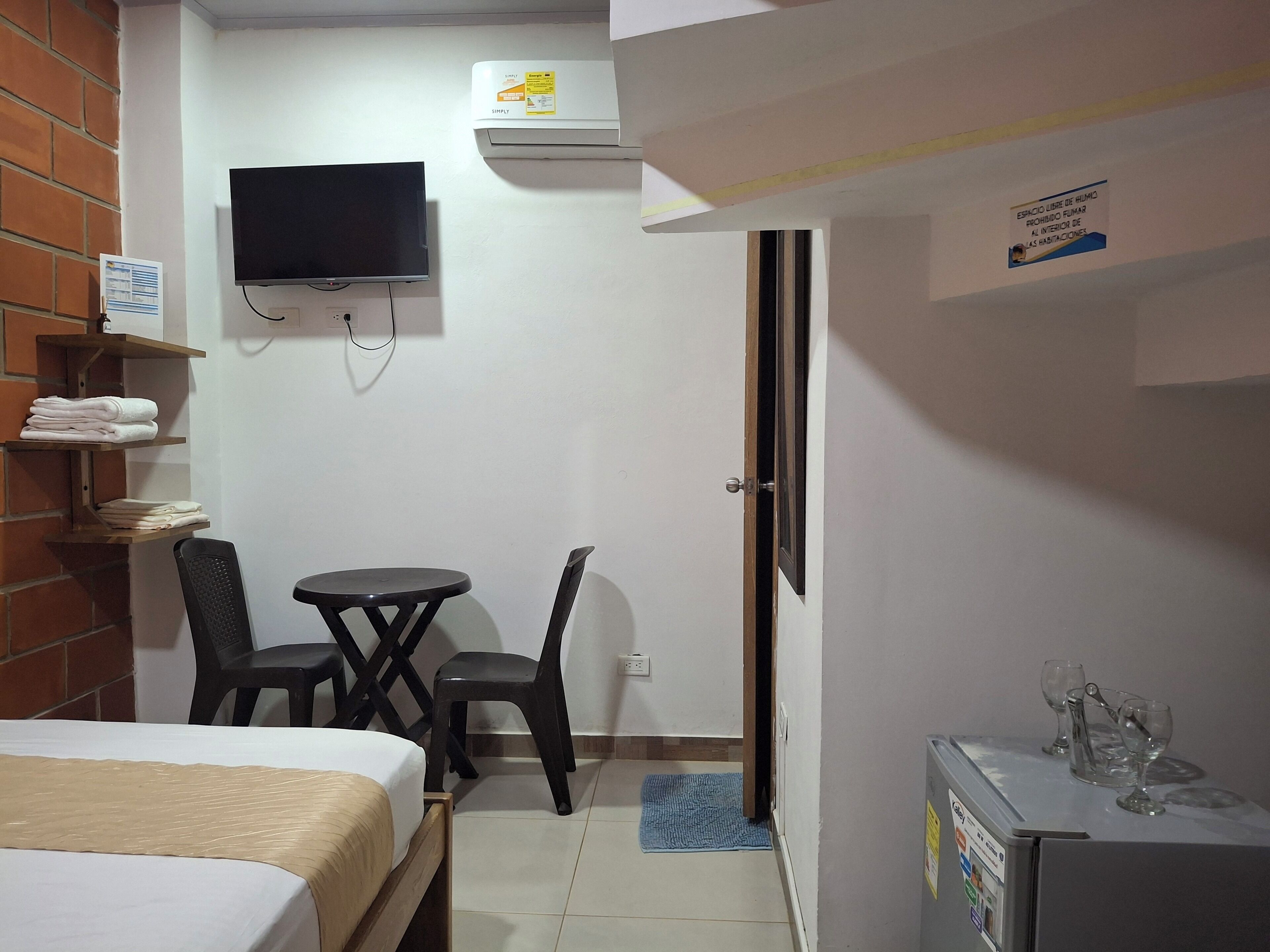 A small hotel room with a bed, a TV mounted on the wall, a desk with a chair, and a refrigerator with wine glasses on top.