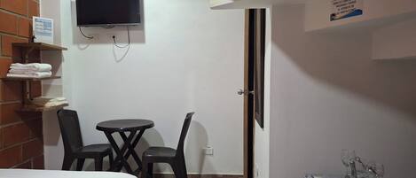 A small hotel room with a bed, a TV mounted on the wall, a desk with a chair, and a refrigerator with wine glasses on top.