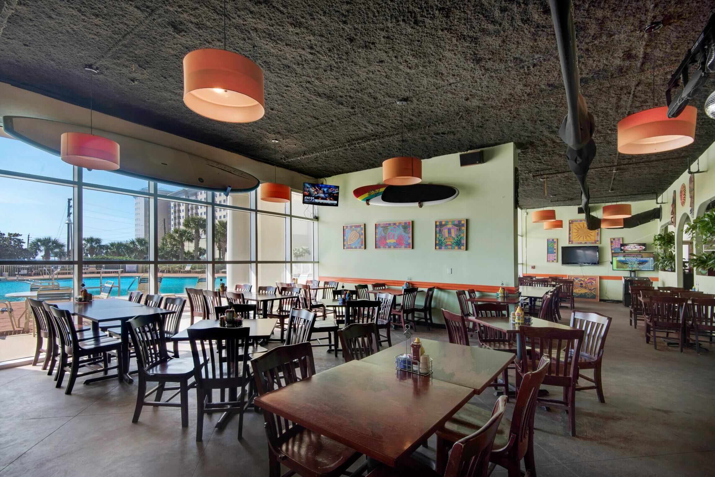 Uma sala de jantar ampla e interna, com grandes janelas, mesas de madeira e luminárias pendentes laranja.
