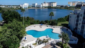 A swimming pool surrounded by a white fence, with a view of a lake and buildings in the background. - INTERNATIONAL dr/UNIVERSAL 2 Bedroom CONDO-HOTEL (Orlando)