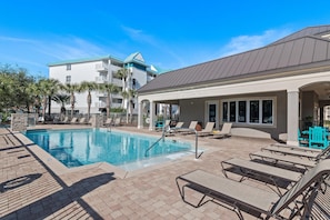 A swimming pool with lounge chairs and a covered area, surrounded by palm trees and a building with a blue roof.