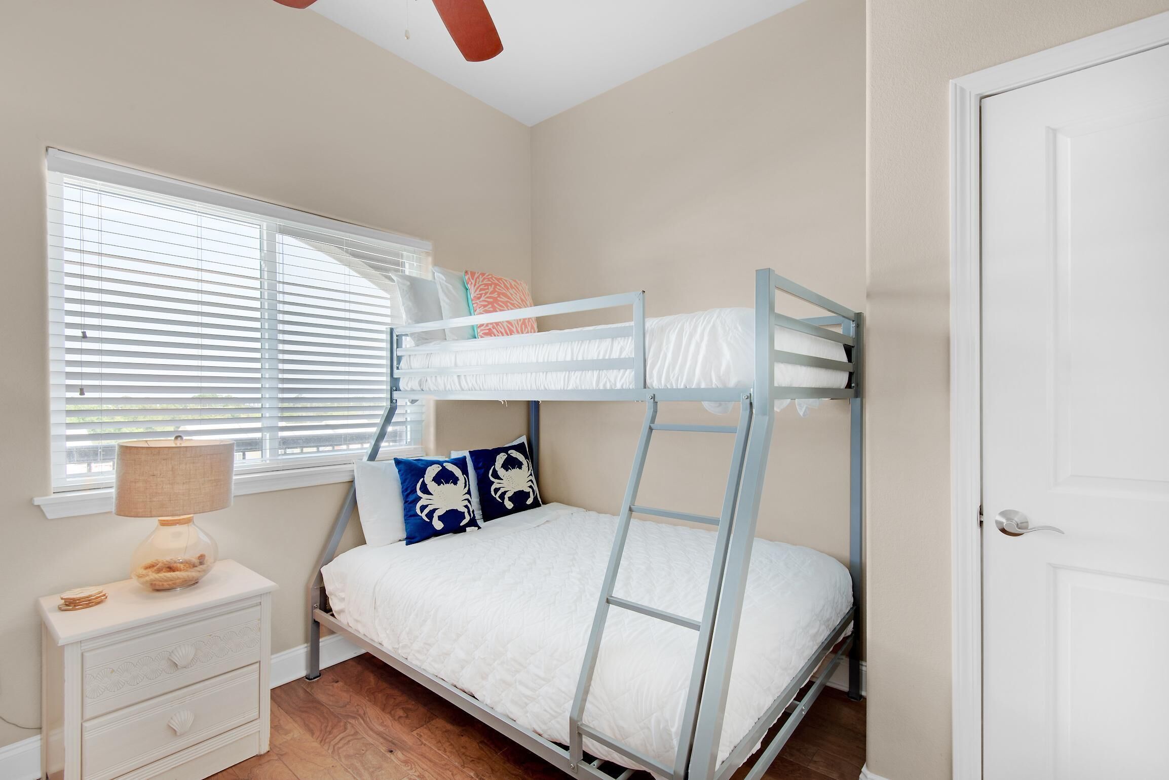 A bunk bed in a room with a window, a bedside table with a lamp, and a door.