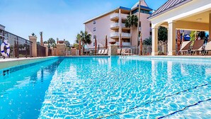 Una piscina con agua azul cristalina, rodeada por un edificio con balcones y una zona cubierta con sillones de descanso.