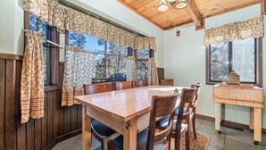 Uma área de refeições com mesa e cadeiras de madeira, janelas amplas com cortinas florais e teto com vigas e luminárias de madeira.
