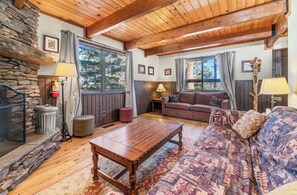 A cozy living room with a stone wall, wooden ceiling beams, a large window, a sofa, a coffee table, and a fireplace.