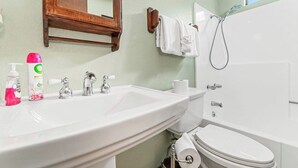 Casa de banho com lavatório branco, sanita e banheira. Há artigos de higiene pessoal no lavatório, um porta-toalhas e um chuveiro.