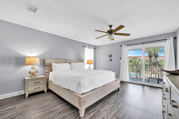 A bedroom with a wooden bed, nightstands, a ceiling fan, and a balcony with a view of palm trees.