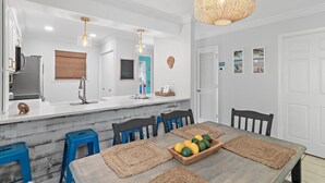 A modern kitchen with white cabinetry, a stainless steel refrigerator, and a blue countertop. There are two blue stools and a wooden table with a fruit bowl. - Summerhouse Townhomes #5 (Miramar Beach)