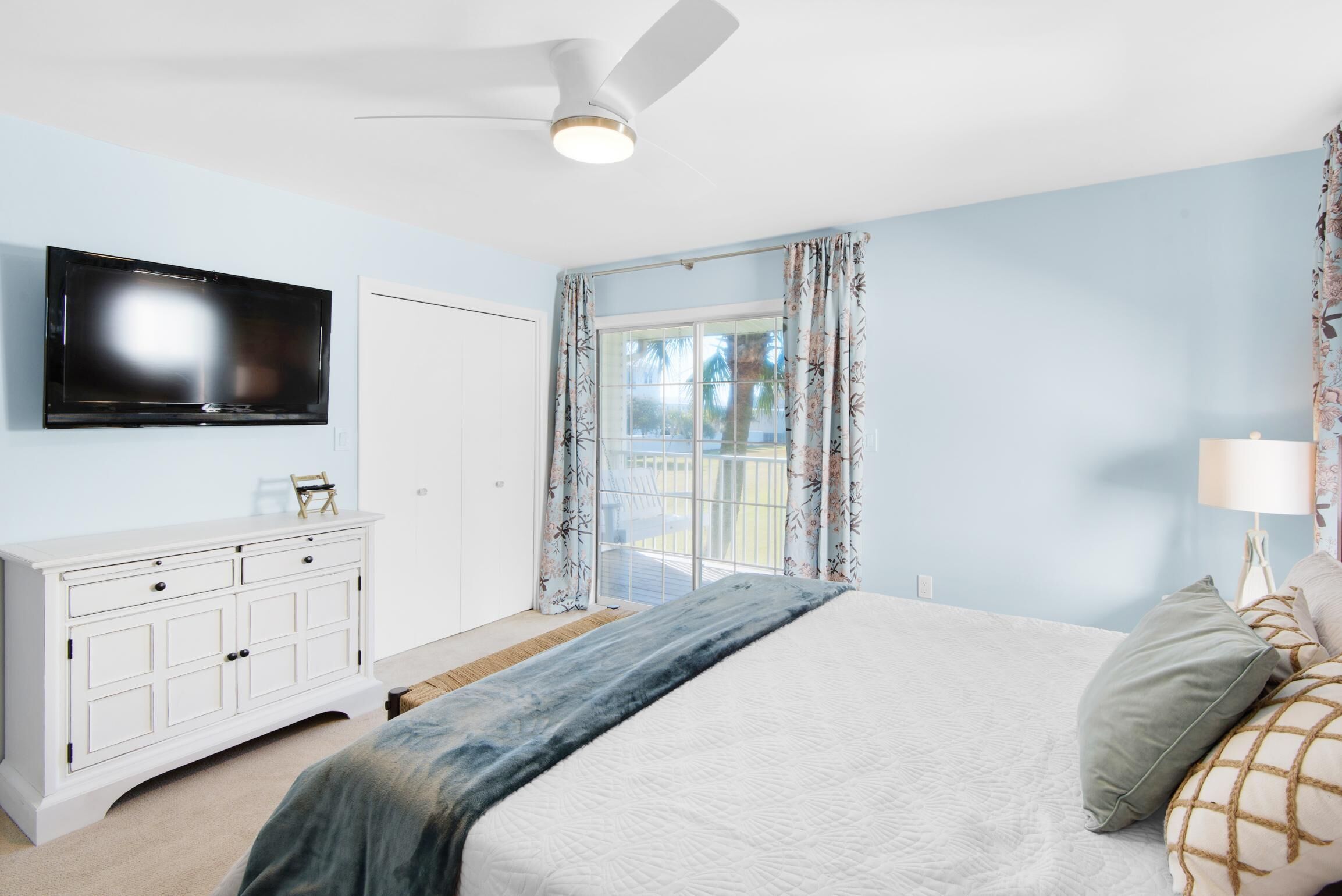 Ein Schlafzimmer mit einem Bett, einem Fernseher, einem Deckenventilator, einem Fenster mit Vorhängen und einer weißen Kommode.