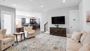 A spacious living room with a large TV, comfortable seating, a patterned rug, and a view into the kitchen area. - Stay Anchored (Miramar Beach)