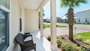 A covered porch with a wicker chair, a palm tree, and a view of a residential street. - Stay Anchored (Miramar Beach)