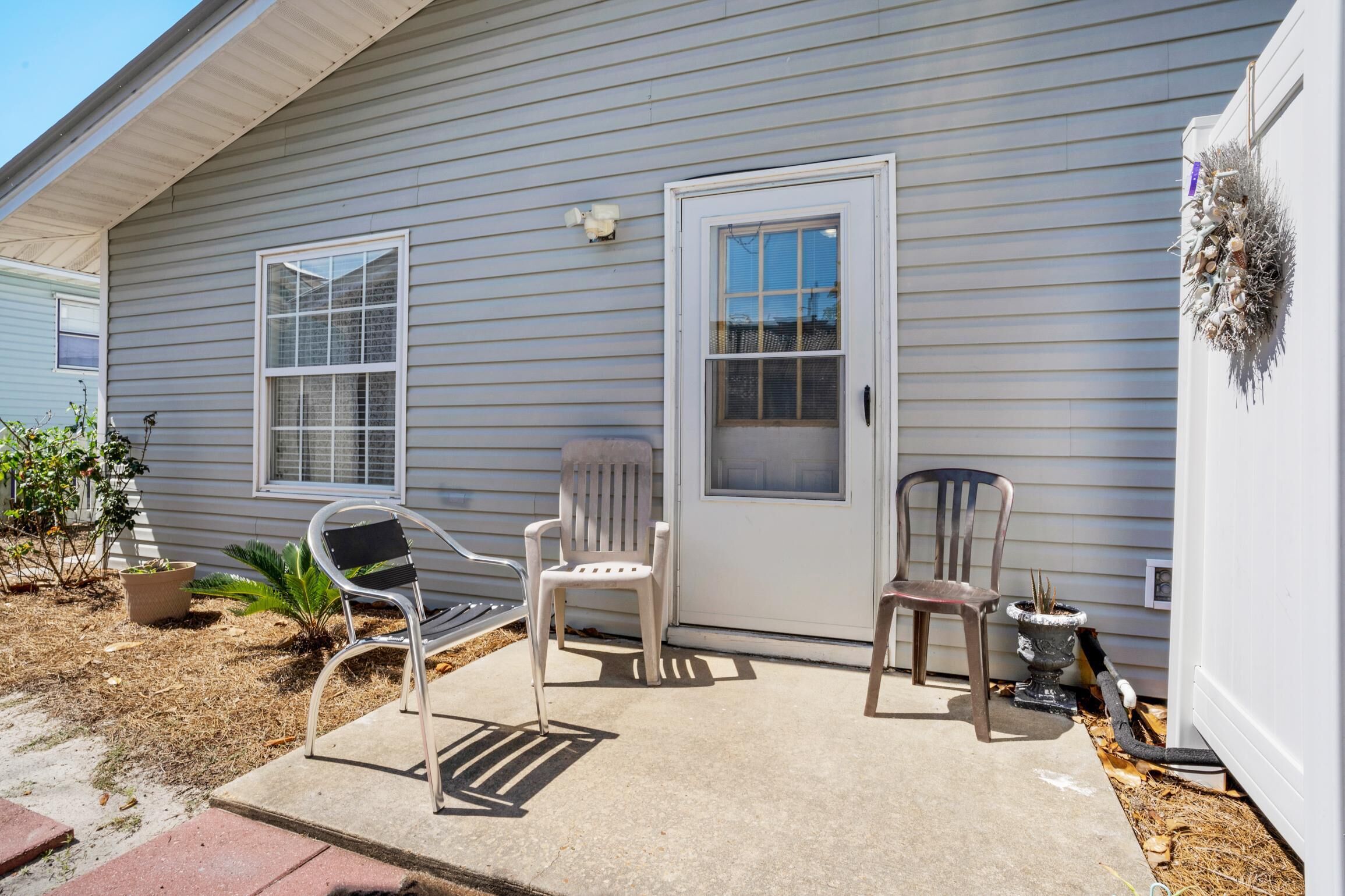 A house with a grey exterior, a front door, two windows, and a concrete patio with two chairs and a potted plant.