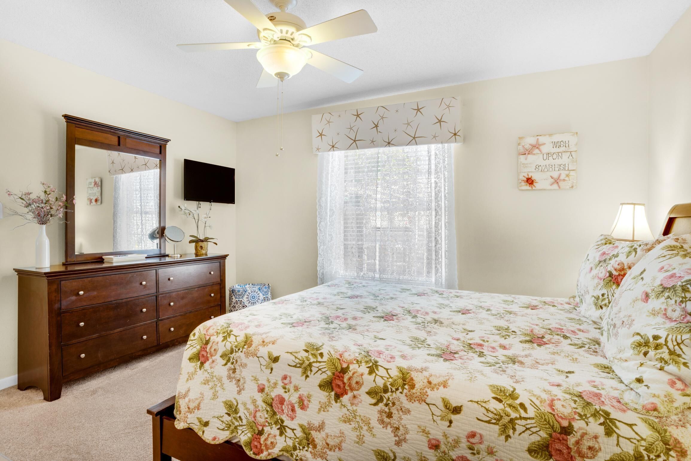 A bedroom with a bed, a dresser, a mirror, a ceiling fan, a window with curtains, and a wall decoration.