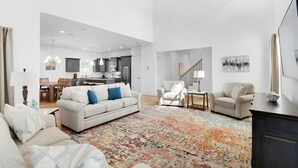 A spacious living room with a large sofa, two armchairs, a coffee table, and a colorful rug. A staircase leads to an upper level with a kitchen and dining area. - Shore Enough (Miramar Beach)