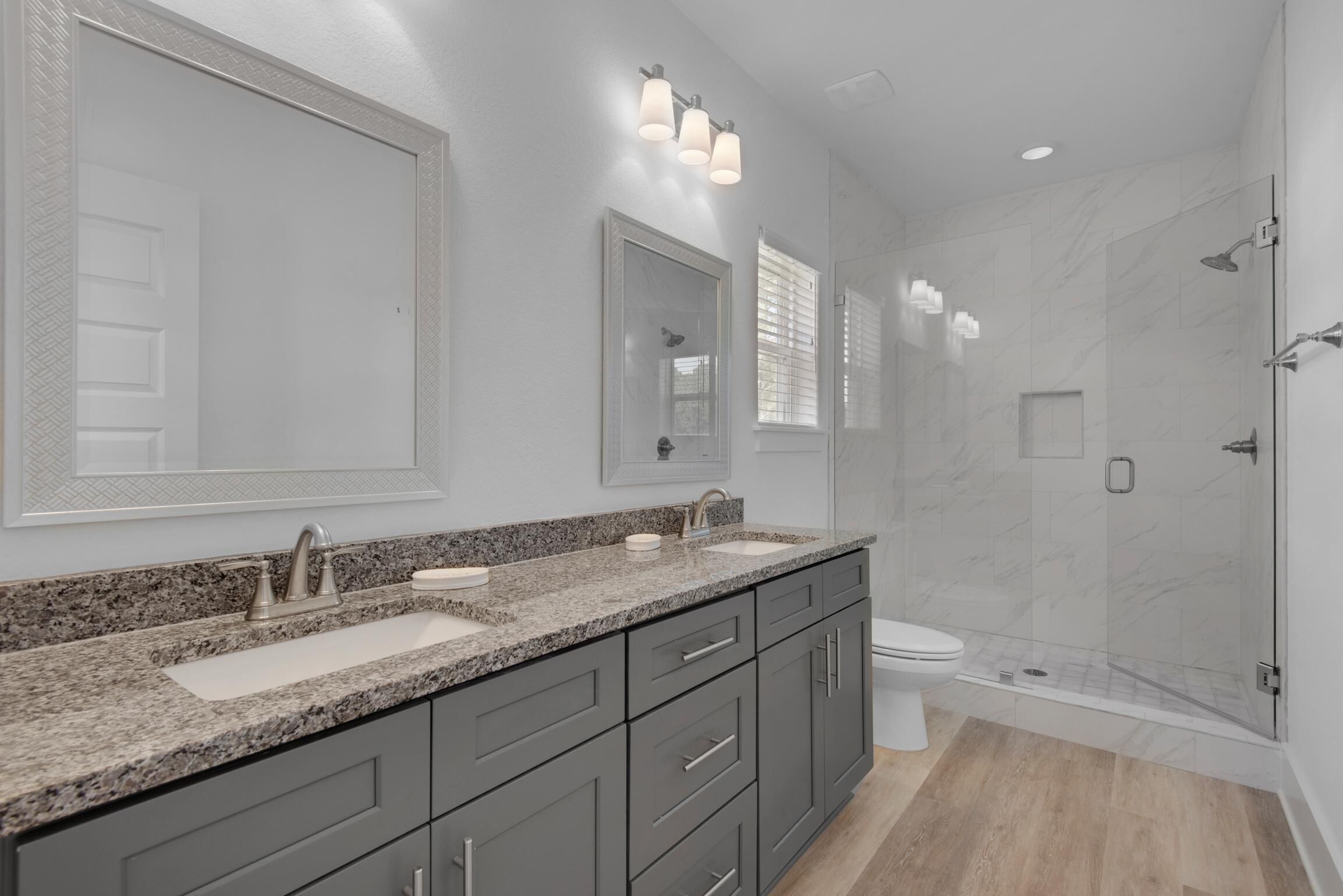 Une salle de bain moderne dotée d’un comptoir en granit, d’un lavabo double, de toilettes et d’une douche à l’italienne.