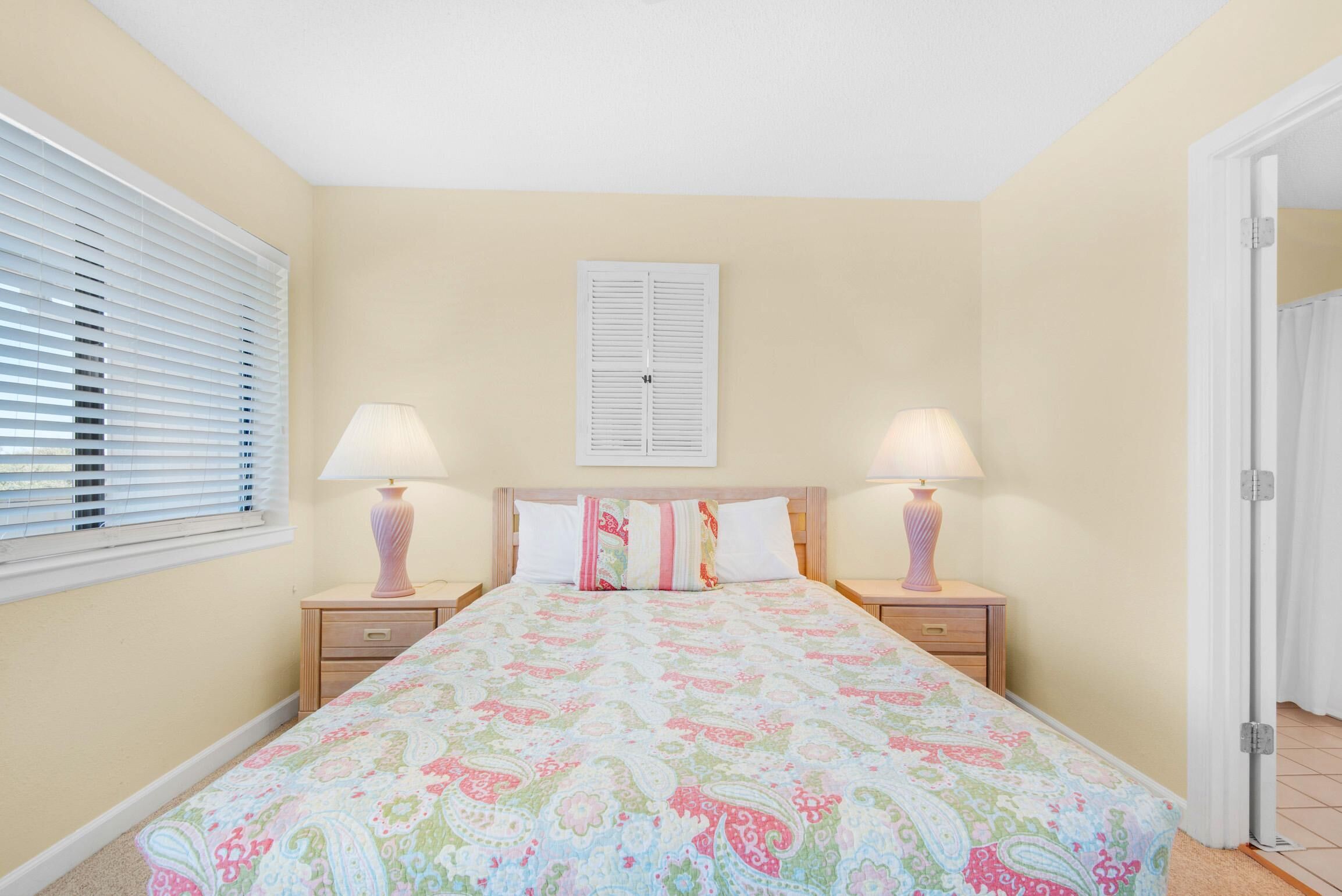 A bedroom with a bed, two bedside tables with lamps, a window with blinds, and a door.
