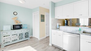 A kitchen with white cabinets, a microwave, and a dishwasher. There is a clock on the wall and a sign that says "The sun and the sand and a drink in my hand."