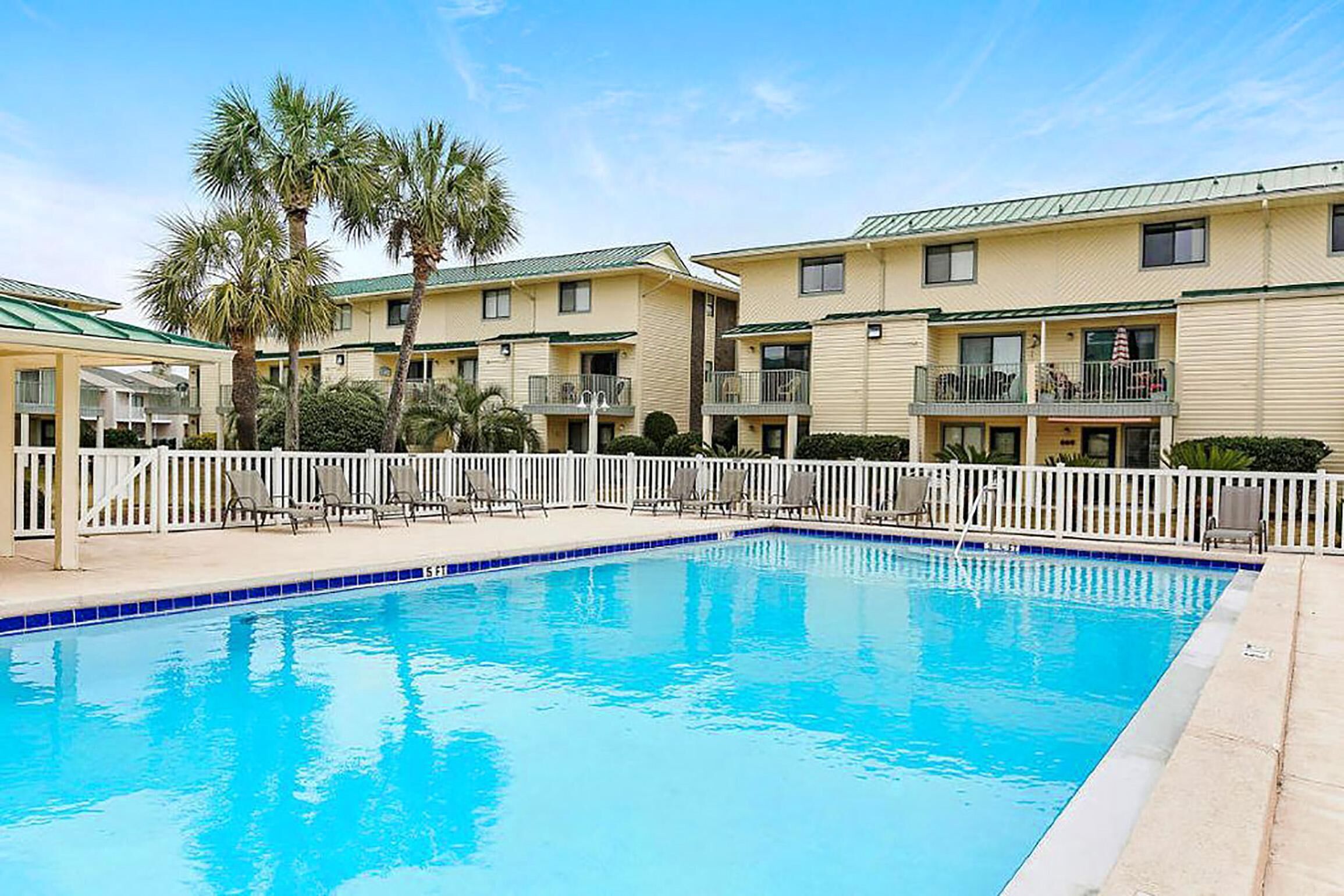 A swimming pool surrounded by a white fence, with a row of two-story buildings in the background.