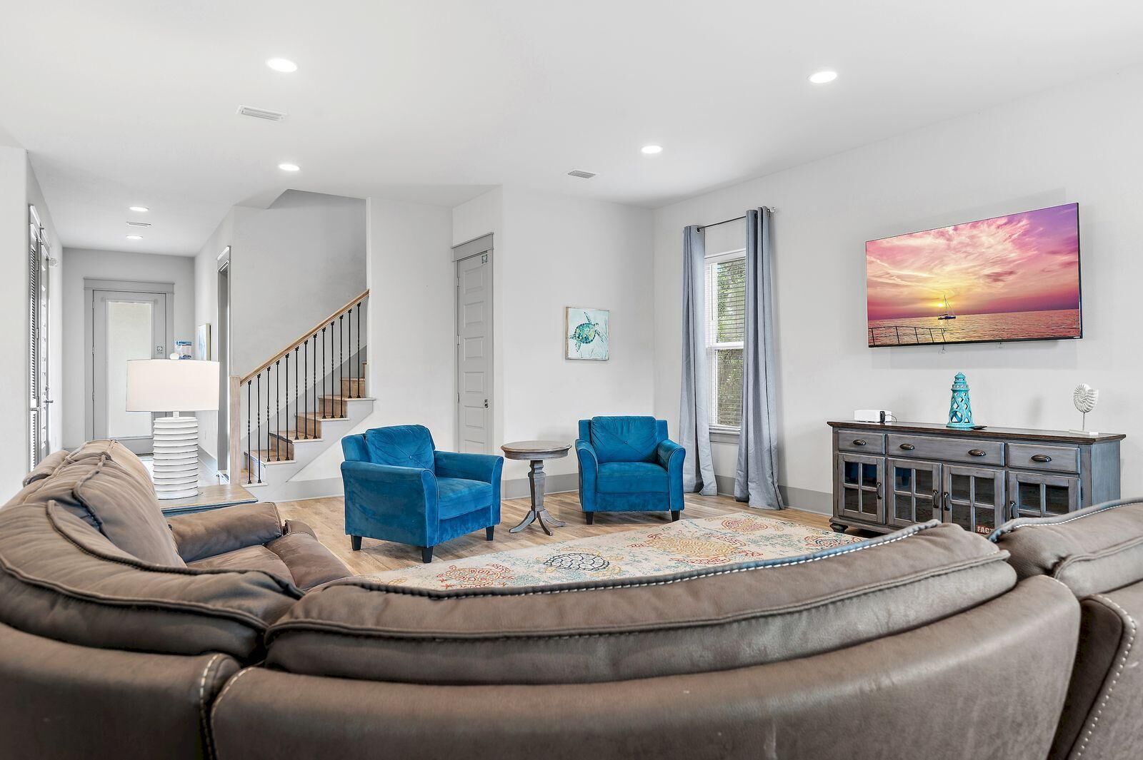 A living room with a brown leather sofa, two blue armchairs, a patterned rug, a cabinet with decorative items, and a sunset painting on the wall.
