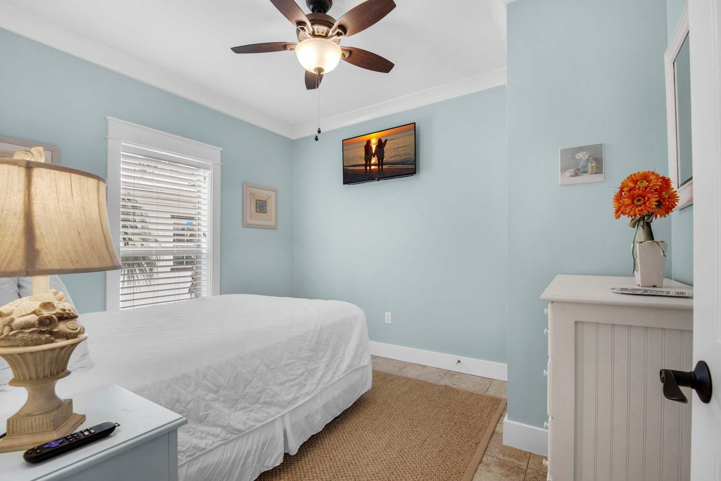 Une chambre à coucher comprenant un lit, un ventilateur de plafond, une télévision, une lampe, une commode et une fenêtre avec des stores.