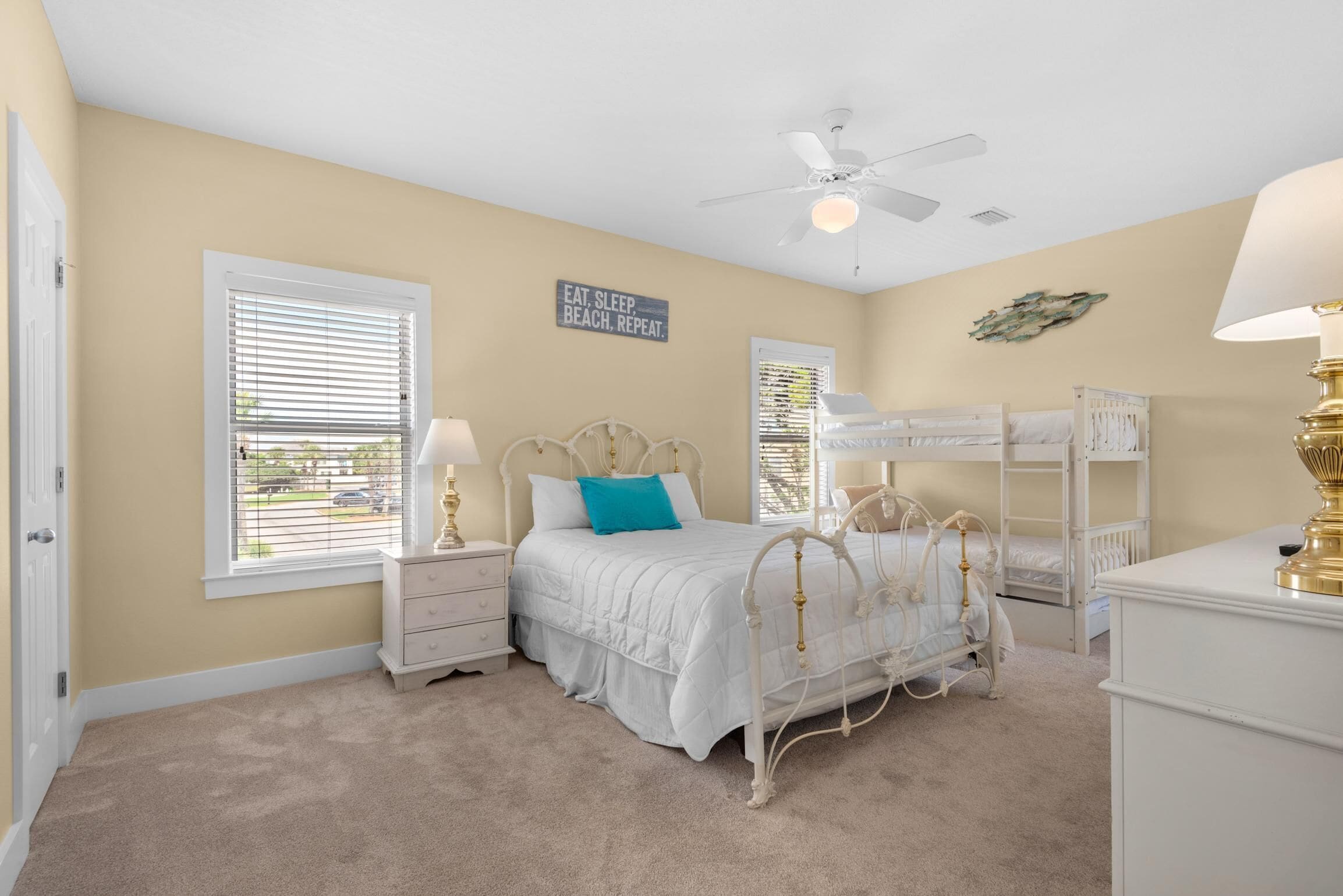 A bedroom with a bunk bed, a single bed, a nightstand, a lamp, a ceiling fan, and a window with blinds.