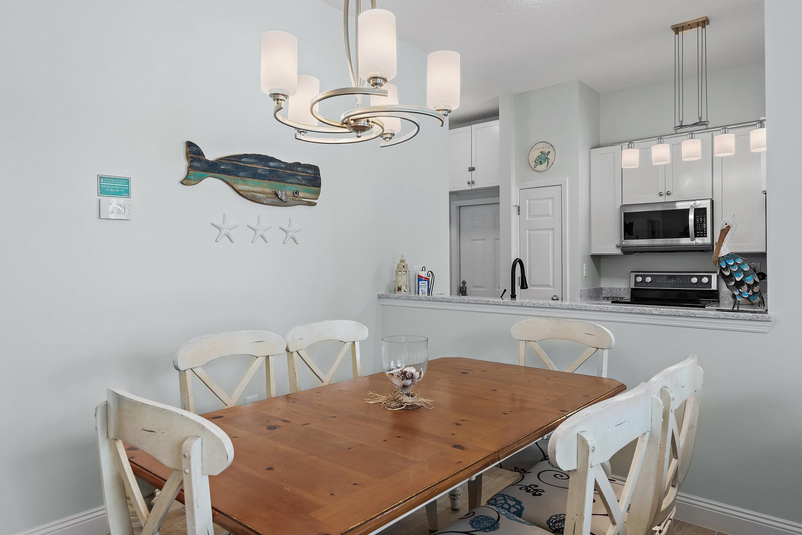 A dining area with a wooden table and white chairs, a chandelier, and a kitchen with white cabinets and a microwave.