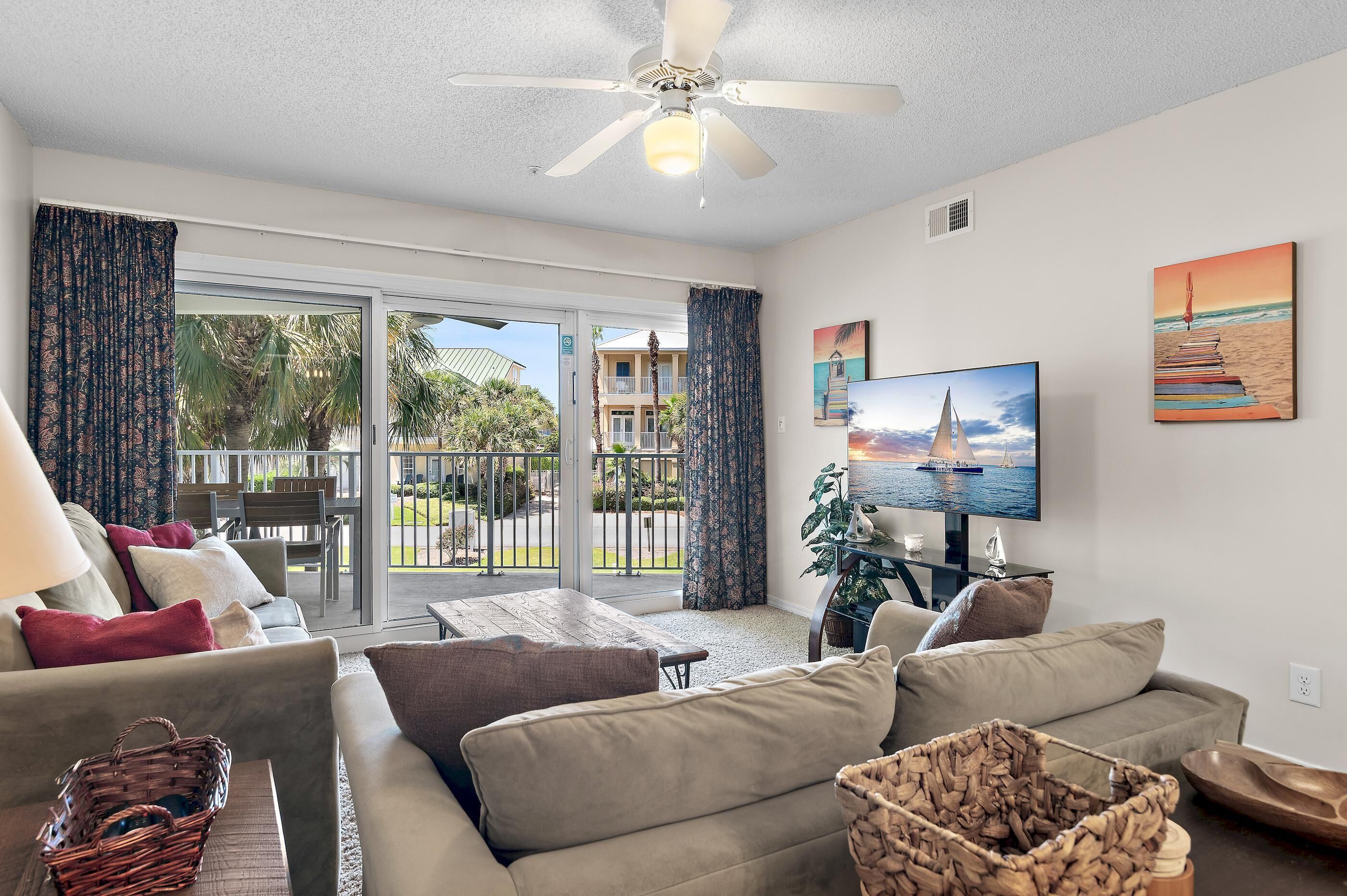 A living room with a sofa, a TV, and a balcony with a view of palm trees and a building.