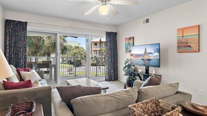 A living room with a sofa, a TV, and a balcony with a view of palm trees and a building. - Maravilla 3202 (Miramar Beach)