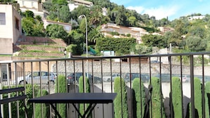 Un balcon avec une table et deux chaises, donnant sur un parking où sont garées des voitures et bordé d’un mur en pierre.