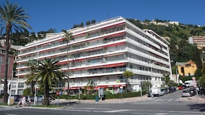 Un hôtel à plusieurs étages, doté de balcons, situé dans une rue ensoleillée bordée de palmiers et où sont garées des voitures.