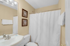 A bathroom with a white sink, mirror, and toilet. There are two framed pictures on the wall, and a white shower curtain. - Maravilla 3107 (Miramar Beach)