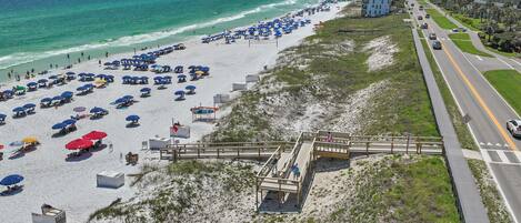 A beach with rows of umbrellas and beach chairs, a wooden boardwalk, and a road parallel to the beach.