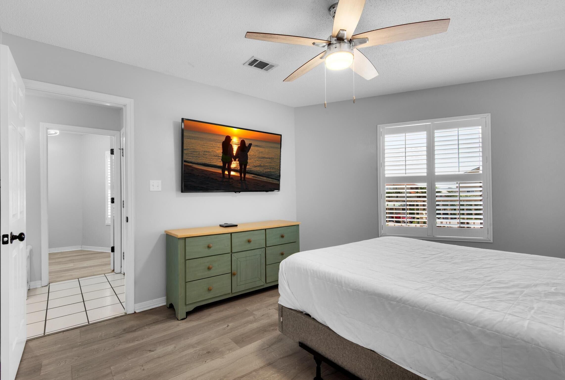 A bedroom with a bed, a ceiling fan, a green dresser, a painting of a sunset, and a window with blinds.