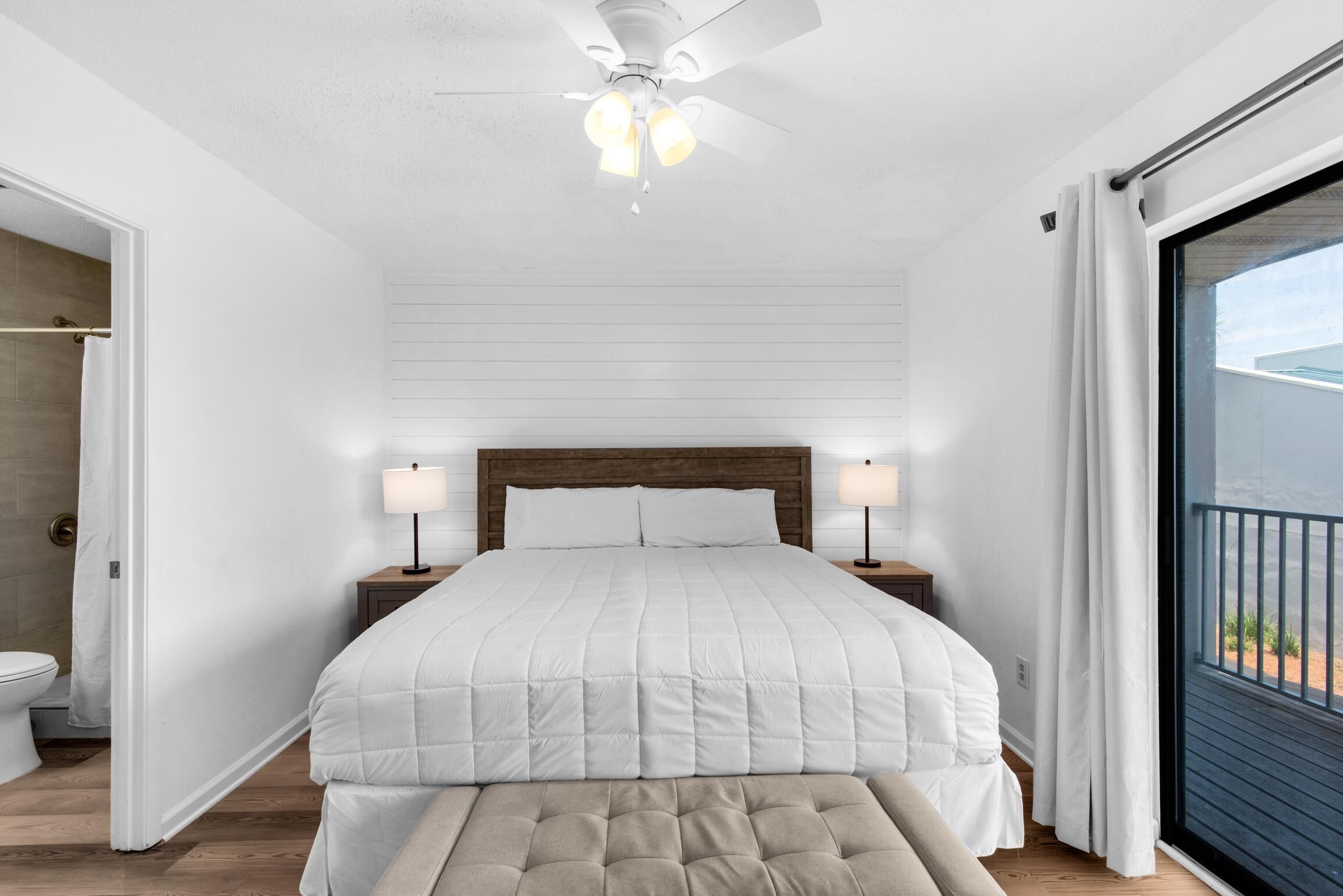 A bedroom with a large bed, two bedside lamps, a ceiling fan, and a view through a sliding glass door.